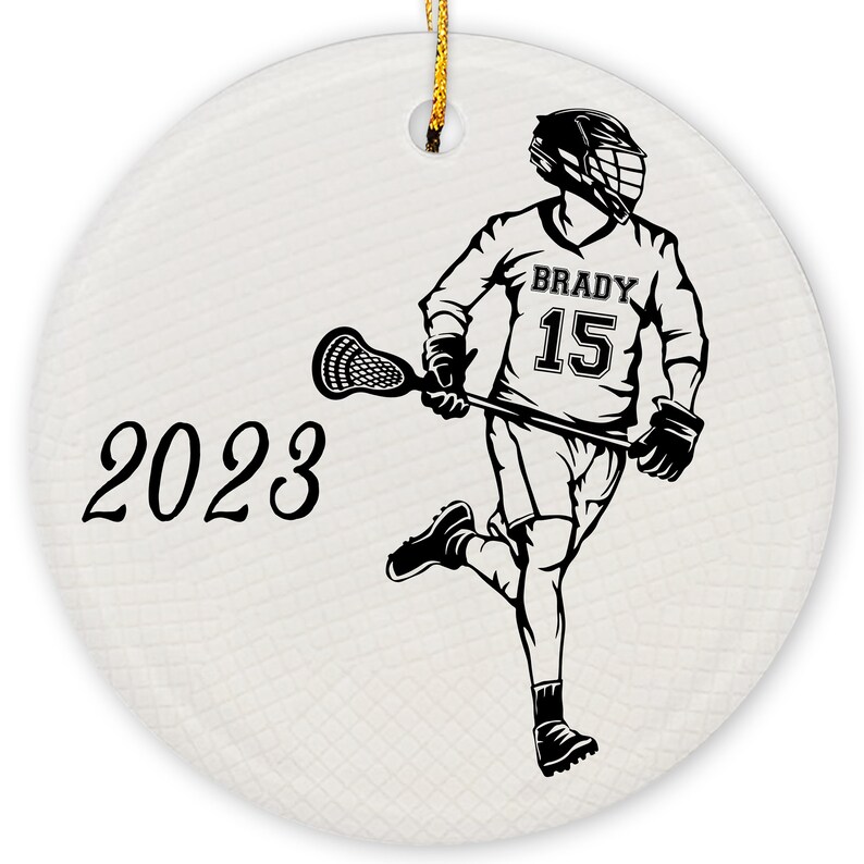 May include: A black and white illustration of a lacrosse player wearing a helmet and jersey with the number 15 and the name "BRADY" on it. The player is holding a lacrosse stick and is running with the ball. The year 2023 is written in black cursive on the left side of the image.