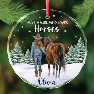 May include: A clear, round ornament with a red ribbon. The ornament features a girl in a denim jacket and cowboy boots, walking with a brown horse in a snowy forest scene. Text reads "JUST A GIRL WHO LOVES Horses" and the name "Olivia".