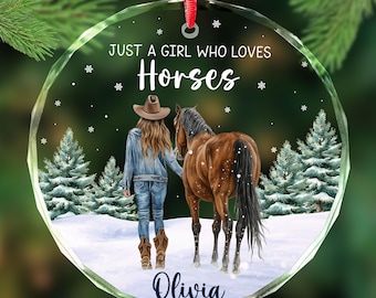 May include: A clear, round Christmas ornament with a red ribbon. It depicts a girl in a denim jacket and cowboy boots, walking with a brown horse in a snowy forest. Text reads "JUST A GIRL WHO LOVES Horses" and the name "Olivia".