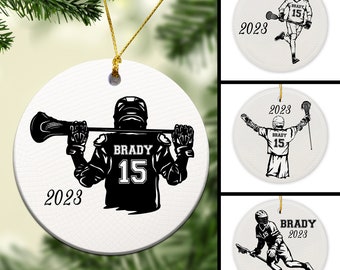 May include: White ceramic ornament with a black silhouette of a lacrosse player holding a lacrosse stick. The player is wearing a jersey with the number 15 and the name "BRADY" on the back. The ornament also includes the year "2023".