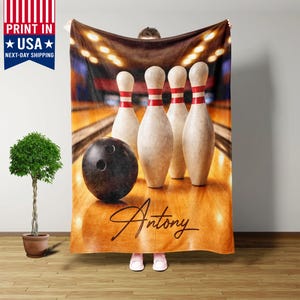 May include: A bowling-themed blanket featuring a black bowling ball and pins with red stripes. The blanket has a wooden lane background and the name "Antony" in cursive. The blanket is held up by a person.