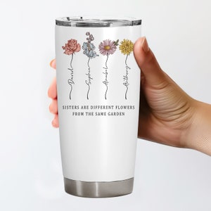 May include: White stainless steel tumbler with a silver base and lid. The tumbler features four colorful flower illustrations with names below each flower. The text "Sisters are different flowers from the same garden" is printed on the tumbler.