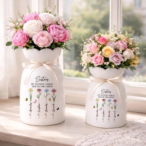 May include: Two white ceramic vases with floral arrangements. The vases have a scalloped top and a ribbon tied around the neck. Text on the vases reads "Sisters are different flowers from the same garden." Each vase has names and flower illustrations.