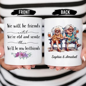 May include: White ceramic mug with a black handle. The front of the mug has a floral design and text that reads "We will be friends until we're old and senile then we'll be new bestfriends". The back of the mug has a cartoon illustration of two older women sitting in chairs with drinks in their hands. The text "Sophia & Amabel" is printed below the illustration.
