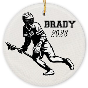May include: Black and white silhouette of a lacrosse player holding a lacrosse stick. The player is wearing a jersey with the number 15. The text "BRADY 2023" is printed on the ornament.