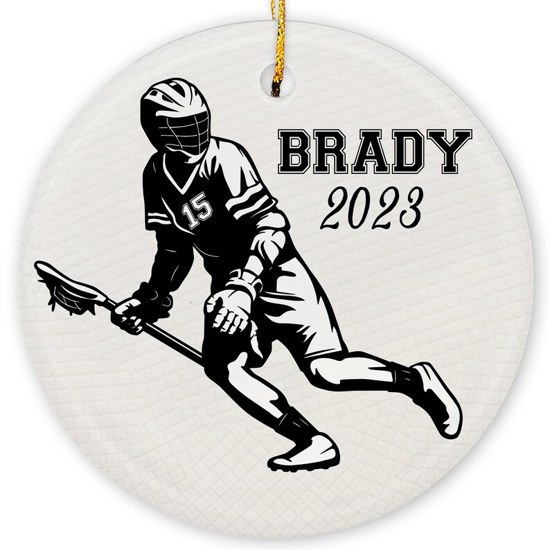 May include: Black and white silhouette of a lacrosse player holding a lacrosse stick. The player is wearing a jersey with the number 15. The text "BRADY 2023" is printed on the ornament.