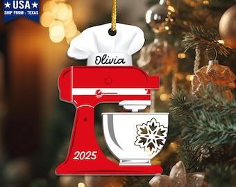 May include: A red and white Christmas ornament shaped like a stand mixer, with the name "Olivia" on a chef's hat. The ornament has a gold string for hanging and the year "2025" on the mixer.