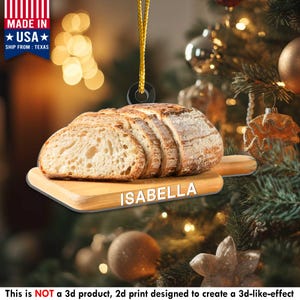 May include: A bread-shaped Christmas ornament with the name "ISABELLA" on a wooden cutting board. The ornament is a 2D print designed to create a 3D-like effect. It has a gold hanging string and is made in the USA.
