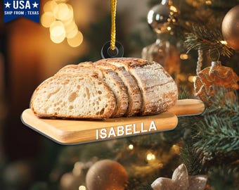 May include: A bread-shaped Christmas ornament with the name "ISABELLA" on a wooden cutting board. The ornament is a 2D print designed to create a 3D-like effect. It has a gold hanging string and is made in the USA.