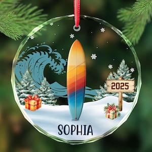 May include: A clear, round Christmas ornament with a surfboard design. The surfboard is vertical, with a gradient of colors from yellow to blue. The ornament includes a wave, snow, trees, gifts, snowflakes, and the name "SOPHIA" at the bottom. A sign reads "2025".