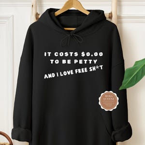 May include: Black hooded sweatshirt with white text: "IT COSTS $0.00 TO BE PETTY AND I LOVE FREE SH*T." A small, round sticker with the text "MOKA QUEENZ" is on the lower right. The sweatshirt is hanging on a wooden rack.