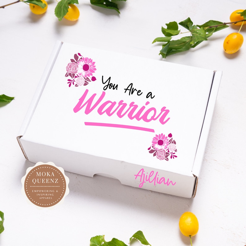 Puede incluir: Caja blanca con detalles florales rosas y el texto "You Are a Warrior". La caja tambi&eacute;n presenta el texto "Ajillian" y un logotipo circular que dice "Moka Queenz Empowering & Inspiring Apparel."