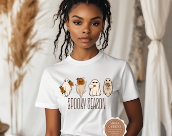 May include: A white short-sleeve t-shirt featuring a graphic of four ghosts with autumn-themed accessories and the text "SPOOKY SEASON" in brown lettering. The ghosts include one holding flowers, one with a pumpkin head, one holding a coffee cup, and one holding a bouquet of dried flowers.