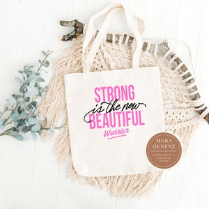 Puede incluir: Un bolso tote de color crema con las palabras "STRONG is the new BEAUTIFUL Warrior" en letras rosas y negras. El bolso tiene asas color crema y se muestra sobre una superficie de madera blanca con elementos decorativos.