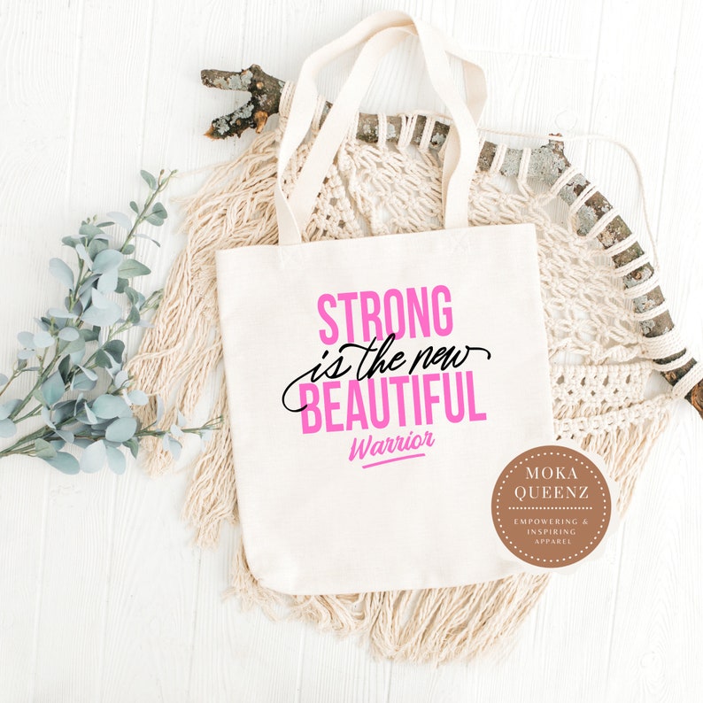 Puede incluir: Un bolso tote de color crema con las palabras "STRONG is the new BEAUTIFUL Warrior" en letras rosas y negras. El bolso tiene asas color crema y se muestra sobre una superficie de madera blanca con elementos decorativos.