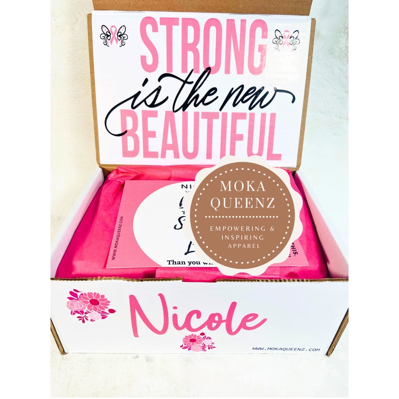 Puede incluir: Una caja blanca con papel de seda rosa en el interior, con el texto "STRONG is the new BEAUTIFUL". La caja tambi&eacute;n muestra el nombre "Nicole" y un logotipo de MOKA QUEENZ, una marca de ropa inspiradora.
