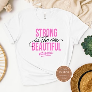 Puede incluir: Camiseta blanca con el texto "STRONG is the new BEAUTIFUL Warrior" en rosa y negro. La camiseta tiene las mangas enrolladas y se muestra sobre un fondo neutro con una suculenta y un sombrero de paja.