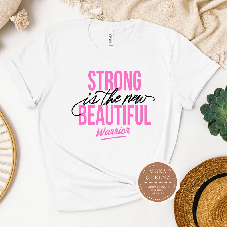 Puede incluir: Camiseta blanca con el texto "STRONG is the new BEAUTIFUL Warrior" en rosa y negro. La camiseta tiene las mangas enrolladas y se muestra sobre un fondo neutro con una suculenta y un sombrero de paja.