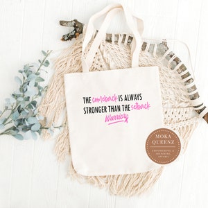 Puede incluir: Bolso tote de color crema con el texto "The comeback is always stronger than the setback Warriors" en negro y rosa. El bolso tiene dos asas y un logotipo redondo que dice "Moka Queenz Empowering & Inspiring Apparel".