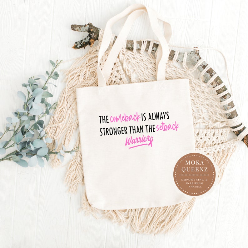 Puede incluir: Bolso tote de color crema con el texto "The comeback is always stronger than the setback Warriors" en negro y rosa. El bolso tiene dos asas y un logotipo redondo que dice "Moka Queenz Empowering & Inspiring Apparel".