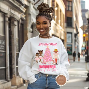 May include: White sweatshirt with a festive graphic featuring a pink Christmas tree, two young girls in tutus, and a nutcracker. The text "It's the Most Wonderful Time of the Year!" is printed in pink.