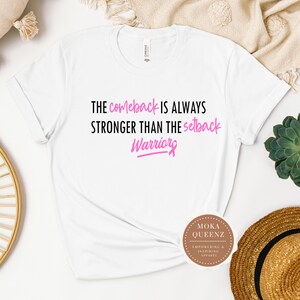Puede incluir: Camiseta blanca con el texto "THE comeback IS ALWAYS STRONGER THAN THE setback Warriors" en negro y rosa. La camiseta tambi&eacute;n presenta un logotipo que dice "MOKA QUEENZ EMPOWERING & INSPIRING APPAREL".