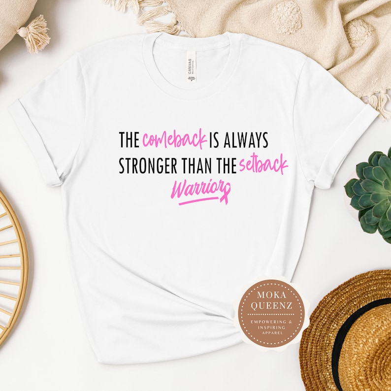 Puede incluir: Camiseta blanca con el texto "THE comeback IS ALWAYS STRONGER THAN THE setback Warriors" en negro y rosa. La camiseta tambi&eacute;n presenta un logotipo que dice "MOKA QUEENZ EMPOWERING & INSPIRING APPAREL".