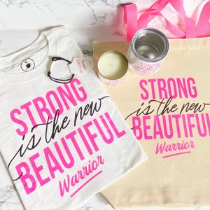 Puede incluir: Conjunto de art&iacute;culos con la frase "Strong is the new Beautiful Warrior" en rosa y negro. Incluye una camiseta blanca, una bolsa de lona, una vela y un vaso. La bolsa tiene un asa rosa.