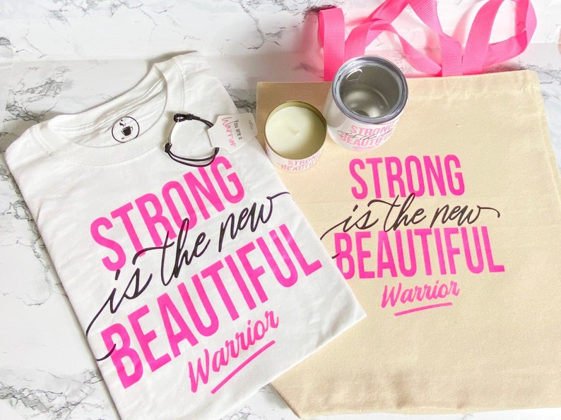 Puede incluir: Conjunto de art&iacute;culos con la frase "Strong is the new Beautiful Warrior" en rosa y negro. Incluye una camiseta blanca, una bolsa de lona, una vela y un vaso. La bolsa tiene un asa rosa.