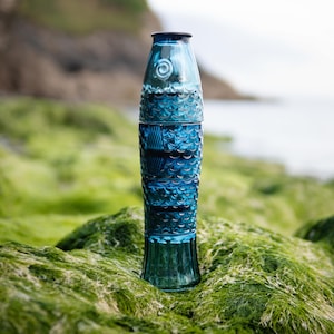 Puede incluir: Una botella alta de vidrio azul con un diseño decorativo. La botella tiene una parte superior azul oscuro y una base acampanada. El diseño incluye patrones texturizados y una espiral. La botella está sobre musgo verde.