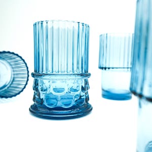 May include: Set of four blue glass tumblers with a ribbed design and a textured base. The glasses are empty and sitting on a white surface.