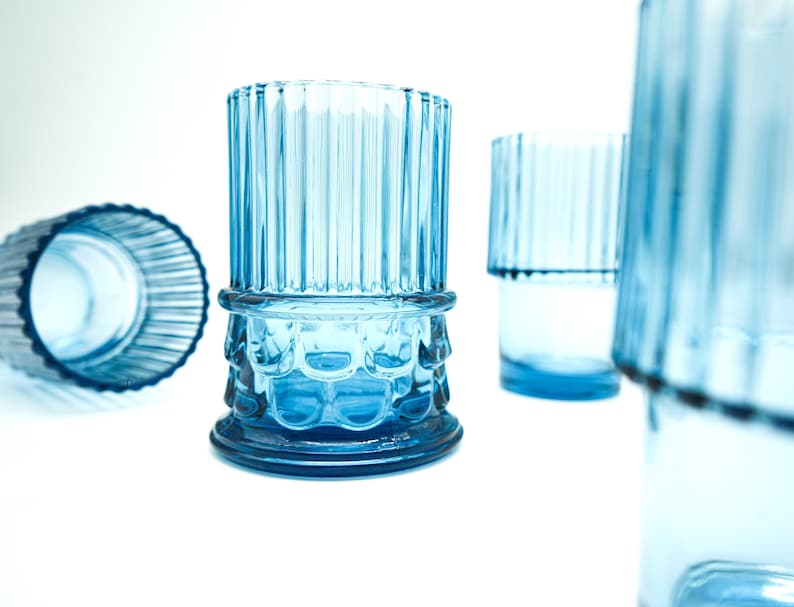 May include: Set of four blue glass tumblers with a ribbed design and a textured base. The glasses are empty and sitting on a white surface.