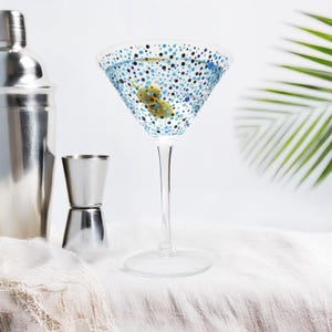 Può includere: Un bicchiere da martini con un motivo a pois blu e neri, riempito con un liquido trasparente e un'oliva verde. Il bicchiere è appoggiato su una tovaglia bianca.