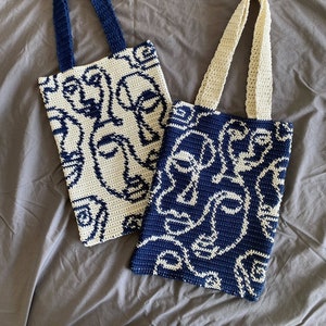 K&ouml;nnte beinhalten: Zwei geh&auml;kelte Einkaufstaschen mit wei&szlig;en und blauen abstrakten Gesichtsdesigns. Die Tasche links hat einen wei&szlig;en Hintergrund mit blauen Gesichtern, und die Tasche rechts hat einen blauen Hintergrund mit wei&szlig;en Gesichtern.