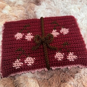 Puede incluir: Una bolsa roja de ganchillo con una cinta marr&oacute;n y dise&ntilde;os de flores rosas. La bolsa tiene un borde festoneado.