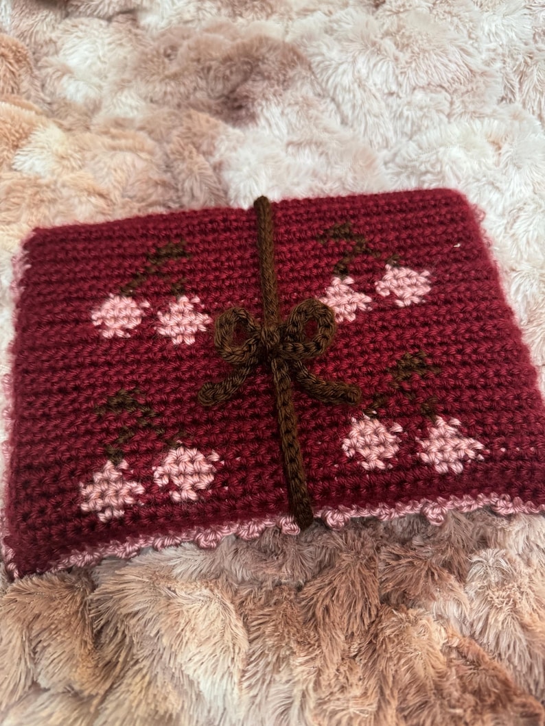 Puede incluir: Una bolsa roja de ganchillo con una cinta marr&oacute;n y dise&ntilde;os de flores rosas. La bolsa tiene un borde festoneado.