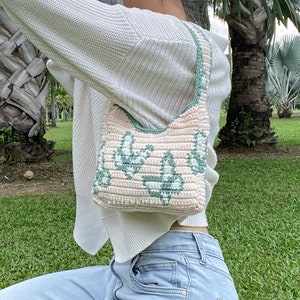May include: A pink and green crocheted handbag with a floral design. The bag is worn over the shoulder of a person wearing a white sweater and blue jeans.