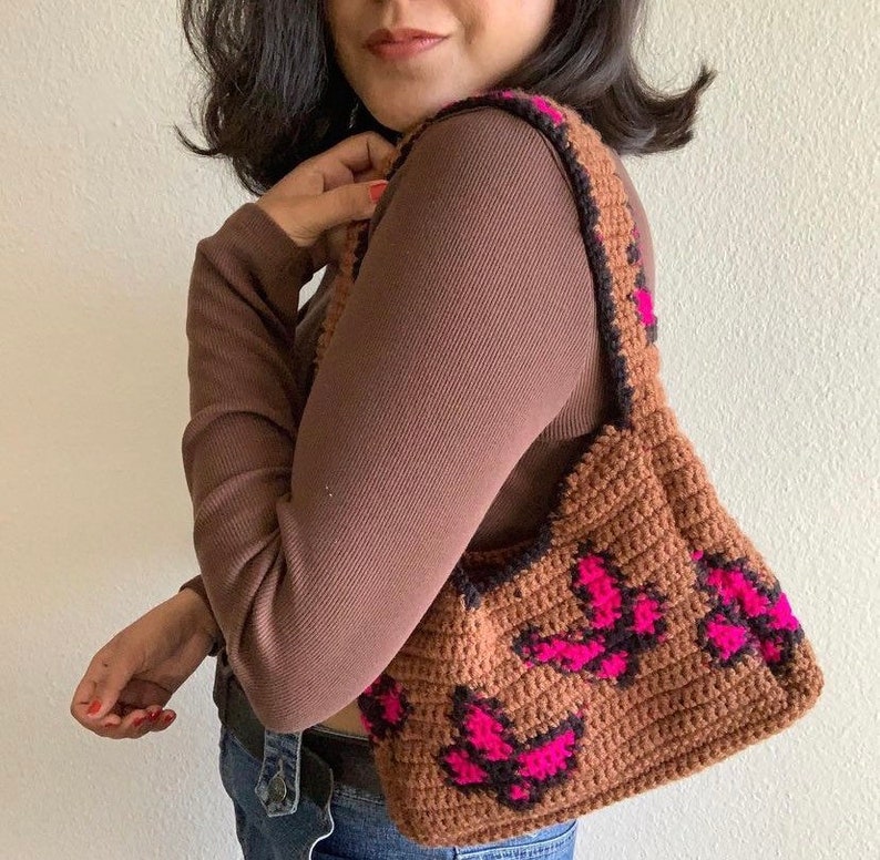 May include: A brown crocheted handbag with a pink and black butterfly design. The bag has a long strap and is worn over the shoulder.