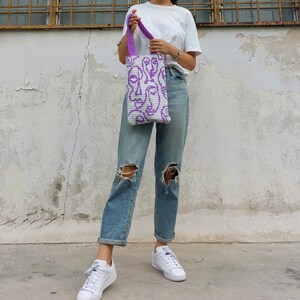 K&ouml;nnte beinhalten: Eine Person tr&auml;gt blaue Jeans und wei&szlig;e Turnschuhe und h&auml;lt eine wei&szlig;e Einkaufstasche mit einem violetten abstrakten Gesichtsdesign.