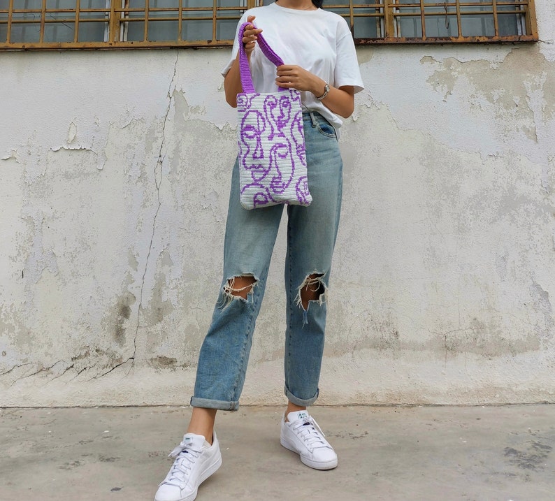 K&ouml;nnte beinhalten: Eine Person tr&auml;gt blaue Jeans und wei&szlig;e Turnschuhe und h&auml;lt eine wei&szlig;e Einkaufstasche mit einem violetten abstrakten Gesichtsdesign.