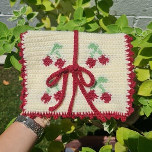 Puede incluir: Una bolsa de ganchillo blanca con un dise&ntilde;o de cerezas rojas y un lazo rojo. La bolsa tiene un borde festoneado.