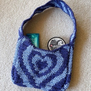 May include: A blue and light blue crocheted tote bag with a rose pattern. The bag is filled with a blue sunscreen bottle and a black jar with the text "One Heck of a Blot".