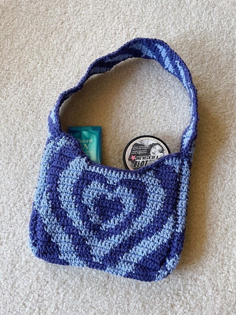 May include: A blue and light blue crocheted tote bag with a rose pattern. The bag is filled with a blue sunscreen bottle and a black jar with the text "One Heck of a Blot".