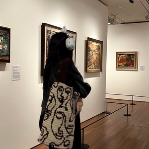 K&ouml;nnte beinhalten: Eine Person in einer schwarzen Lederjacke und einer gestrickten Tragetasche mit einem Gesichtsmuster steht in einer Kunstgalerie. Die Tasche ist cremefarben und braun mit einem sich wiederholenden Muster von Gesichtern. Die Person tr&auml;gt Kopfh&ouml;rer und betrachtet ein Gem&auml;lde an der Wand.