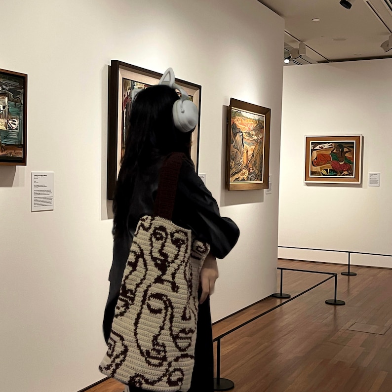 K&ouml;nnte beinhalten: Eine Person in einer schwarzen Lederjacke und einer gestrickten Tragetasche mit einem Gesichtsmuster steht in einer Kunstgalerie. Die Tasche ist cremefarben und braun mit einem sich wiederholenden Muster von Gesichtern. Die Person tr&auml;gt Kopfh&ouml;rer und betrachtet ein Gem&auml;lde an der Wand.