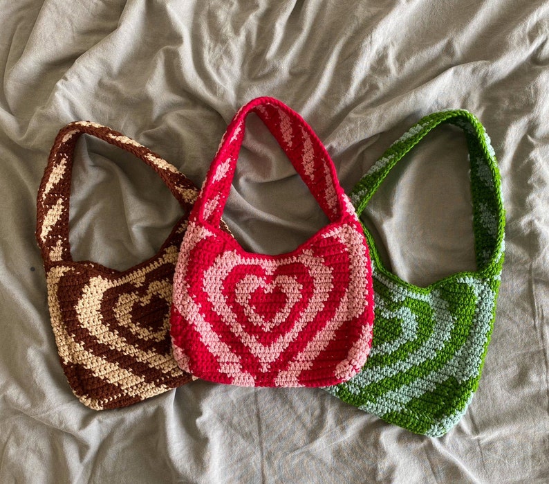 May include: Three crocheted heart-shaped bags in brown, pink, and green. The bags have a single strap and are made with a textured yarn.