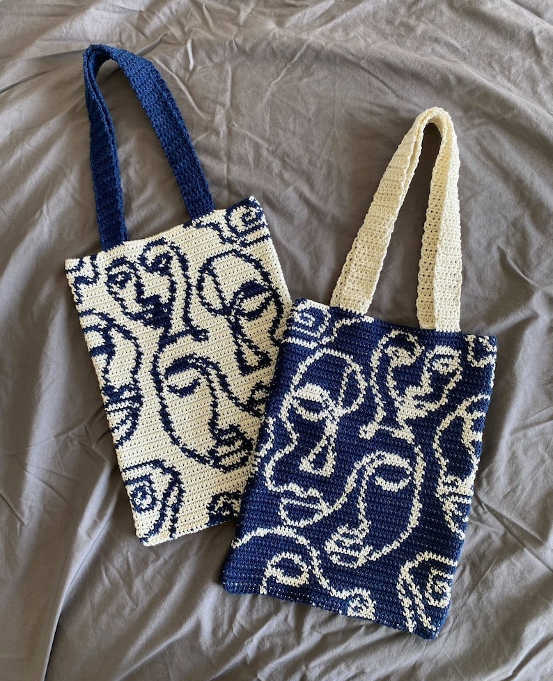 K&ouml;nnte beinhalten: Zwei geh&auml;kelte Einkaufstaschen mit wei&szlig;en und blauen abstrakten Gesichtsdesigns. Die Tasche links hat einen wei&szlig;en Hintergrund mit blauen Gesichtern, und die Tasche rechts hat einen blauen Hintergrund mit wei&szlig;en Gesichtern.