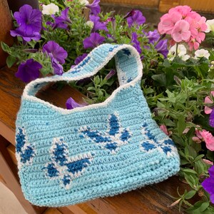 May include: A light blue crocheted bag with a white and blue floral pattern. The bag has a single strap and is made of a soft, textured yarn.