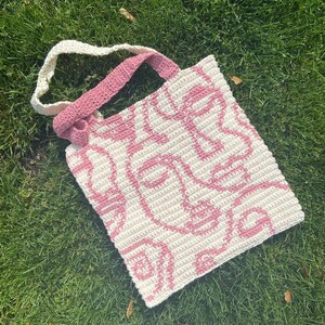 K&ouml;nnte beinhalten: Eine rosa und wei&szlig;e geh&auml;kelte Tragetasche mit einem Muster aus abstrakten Gesichtern. Die Tasche hat einen langen Riemen, der &uuml;ber der Schulter getragen werden kann.