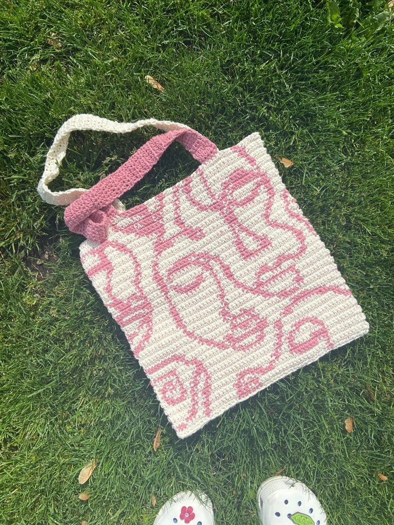 K&ouml;nnte beinhalten: Eine rosa und wei&szlig;e geh&auml;kelte Tragetasche mit einem Muster aus abstrakten Gesichtern. Die Tasche hat einen langen Riemen, der &uuml;ber der Schulter getragen werden kann.